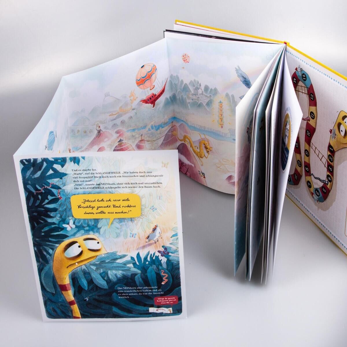 Eine offene Seite aus dem Kinderbuch 'Das NEINhorn und die SchLANGEWEILE'. Die Illustration zeigt eine gelbe Schlange mit müdem Ausdruck in einer blauen Landschaft, während weitere Seiten des Buches Abenteuer und bunte Charaktere darstellen.