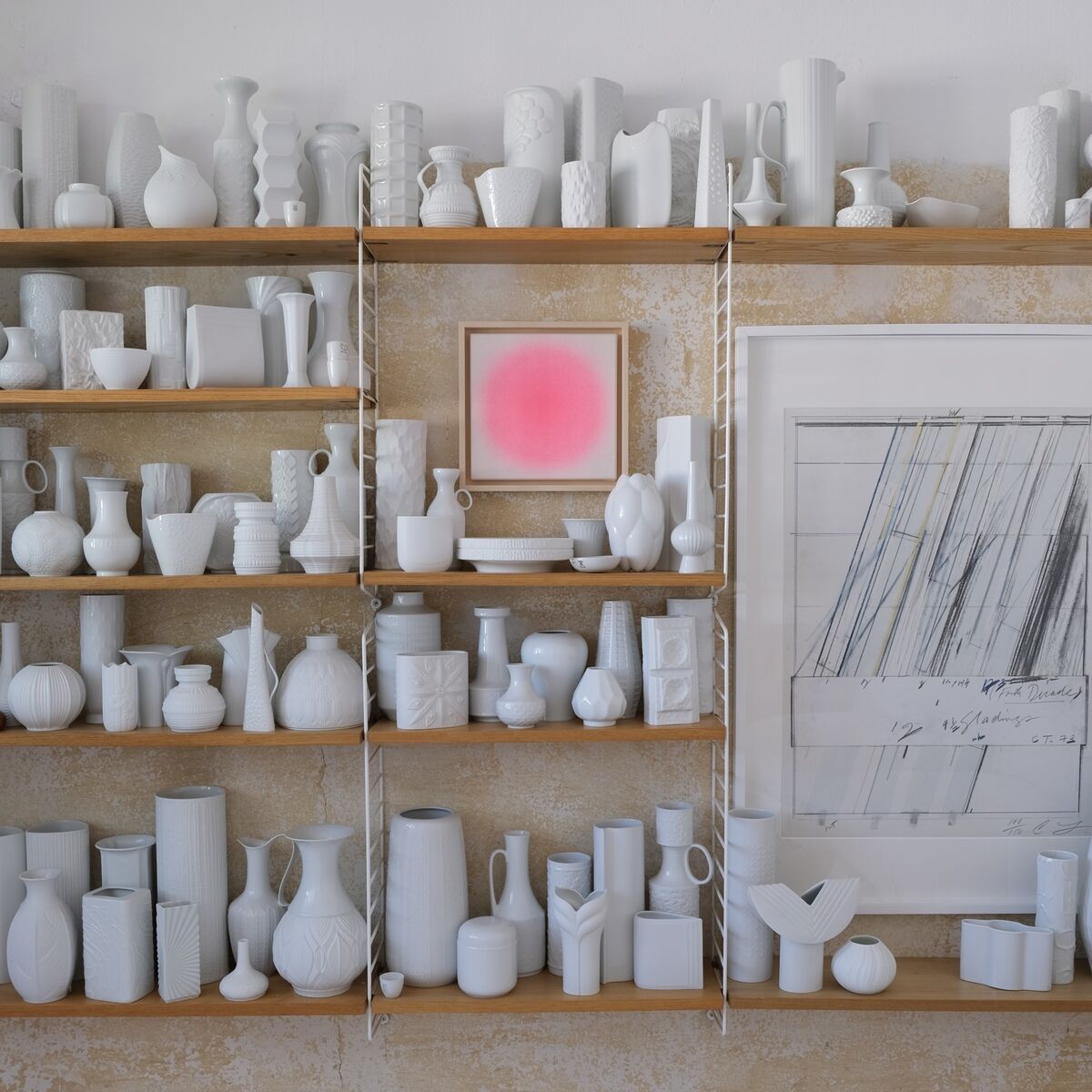 Auf dem Bild ist ein Regal mit zahlreichen weißen Keramikvasen und geometrischen Formen vor einer beigen Wand zu sehen. Im Mittelpunkt hängt ein quadratisches Kunstwerk in leuchtendem Pink der ZEIT-Sonderedition Sorbet. Das Bild bringt einen modernen, farbigen Akzent in die ansonsten zurückhaltend gestaltete Umgebung und verleiht dem Raum eine elegante, zeitgenössische Atmosphäre.