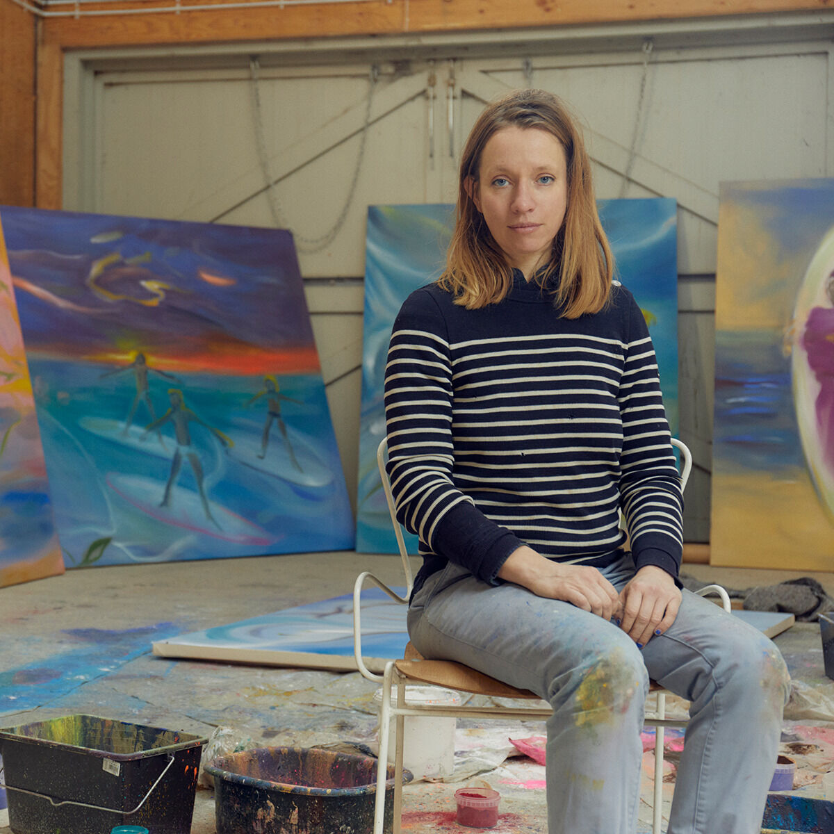 Eine gerahmte Lithografie der ZEIT-Sonderedition »DG 75A« von 2024, präsentiert in einem hell eingerichteten Atelier mit verschiedenen bunten Kunstwerken im Hintergrund. Eine Frau sitzt auf einem Stuhl vor der Kunst und trägt eine gestreifte Pullover.