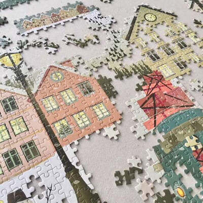 Mehrere zusammengesetzte Puzzleteile zeigen eine verschneite Winterdorfszene mit bunten Häusern, Straßenlaternen und winterlicher Atmosphäre. Das Puzzle liegt auf einem Tisch, einige Teile fehlen noch. Die Illustration vermittelt Weihnachts- und Winterstimmung.