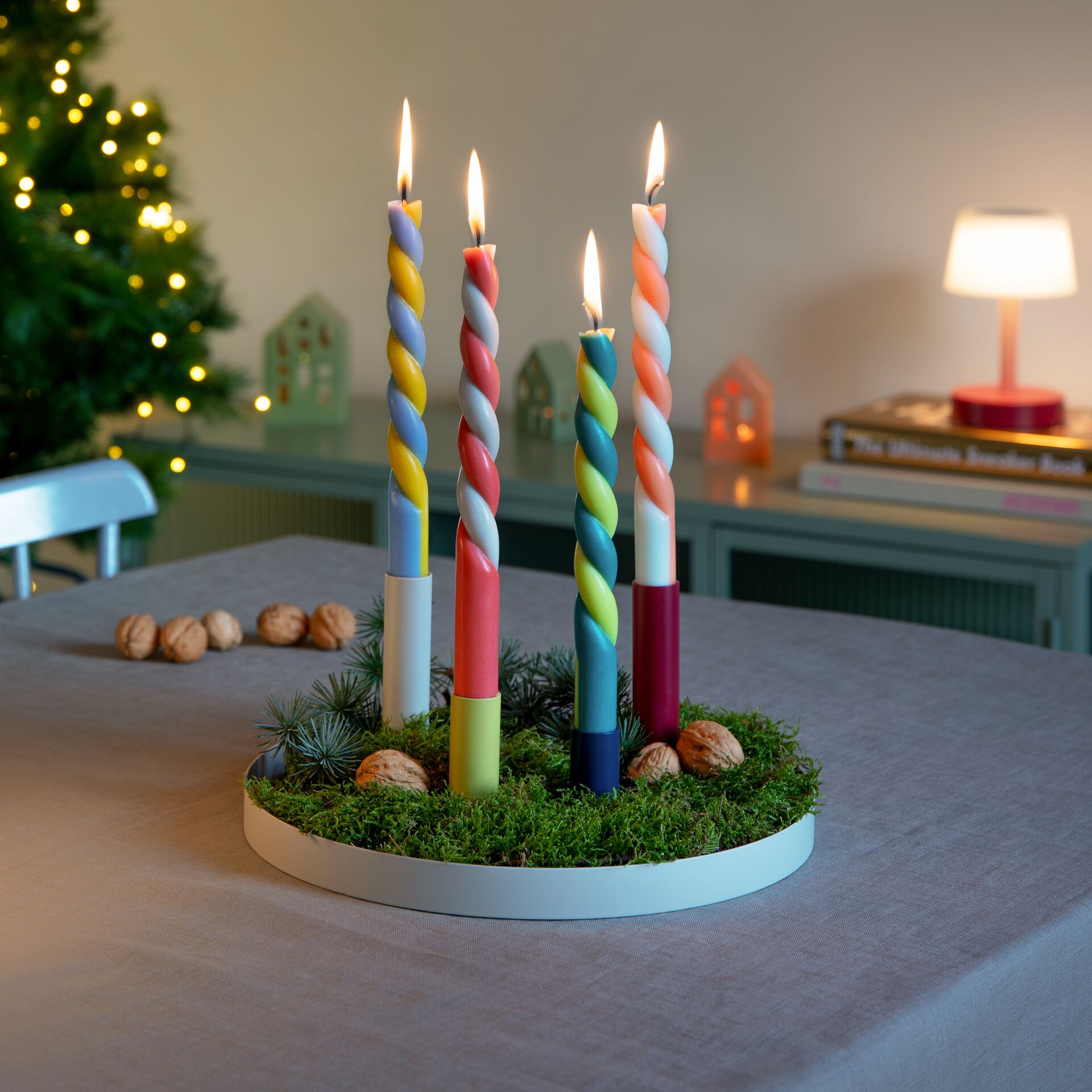 Magnetischer Kerzenhalter 'Luna' mit vier bunten, spiralförmigen Kerzen auf einem weißen Tablett, dekoriert mit Moos und Tannenzweigen, steht als festliche Tischdekoration auf einem Tisch. Im Hintergrund sind Lichter eines Weihnachtsbaums und eine beleuchtete Lampe sichtbar.
