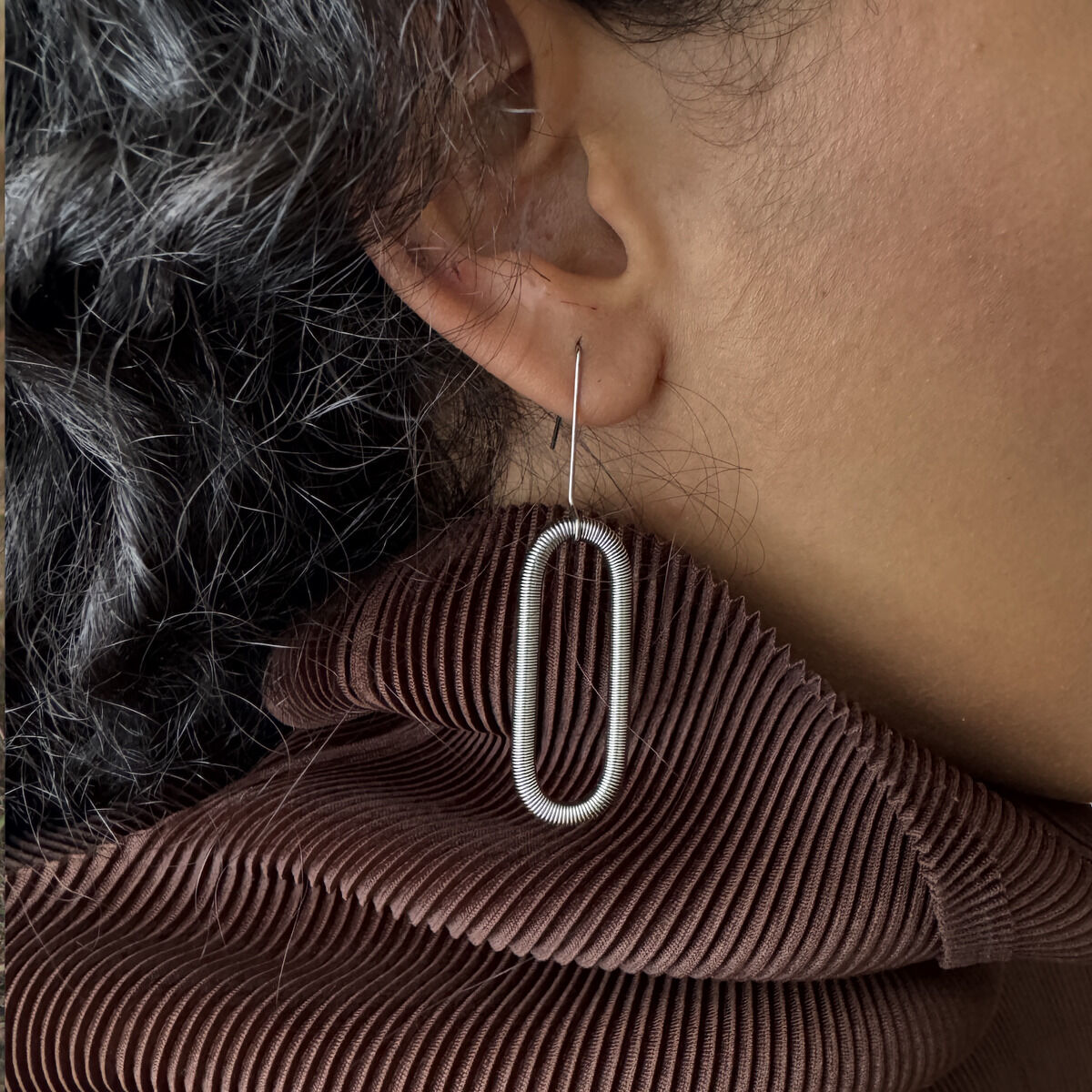 Detailaufnahme eines silbernen Edelstahl-Ohrrings "CHAIN" der Marke la molla von Eolienne Lager am Ohr einer Person. Der Ohrring ist oval, filigran und hängt an einem Ohrläppchen. Im Hintergrund ist eine braune, plissierte Stoffoberfläche sichtbar.