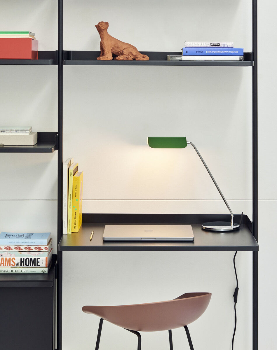 Ein modern eingerichteter Arbeitsplatz mit einer stilvollen Schreibtischlampe aus dem 'Apex Lampe »Desk«' Sortiment. Auf dem Schreibtisch befindet sich ein Laptop, umgeben von stapelbaren Büchern. Ein Trittsessel ist im Vordergrund und zwei Regale sind aufgebaut, eines mit Büchern und eines mit einer braunen Figur darauf.