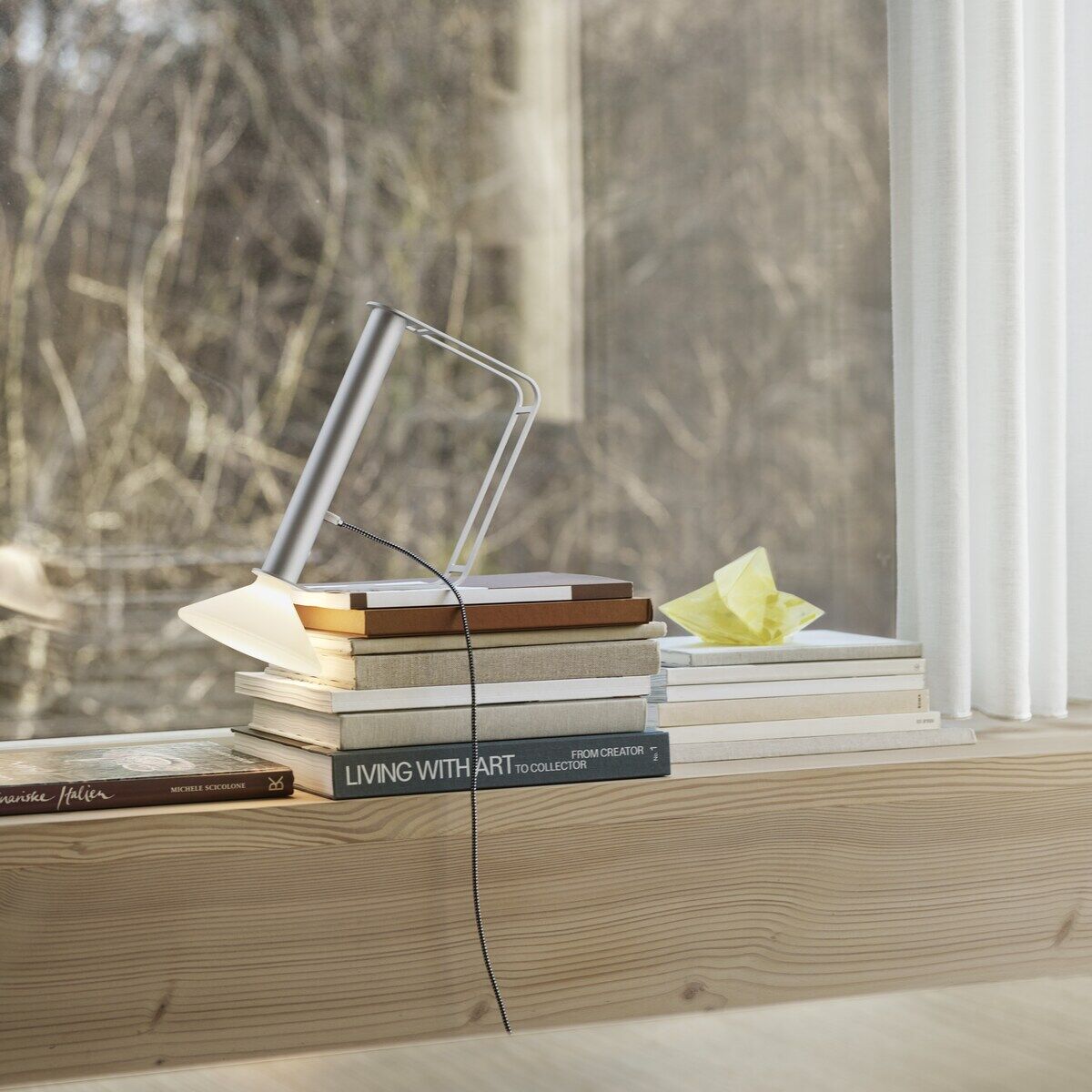 Eine tragbare Lampe »Piton« steht auf einem Holztisch, umgeben von einem Stapel Bücher, vor einem Fenster mit einem Wald im Hintergrund. Die Lampe hat ein minimalistisches, modernes Design mit einem silbernen Körper.