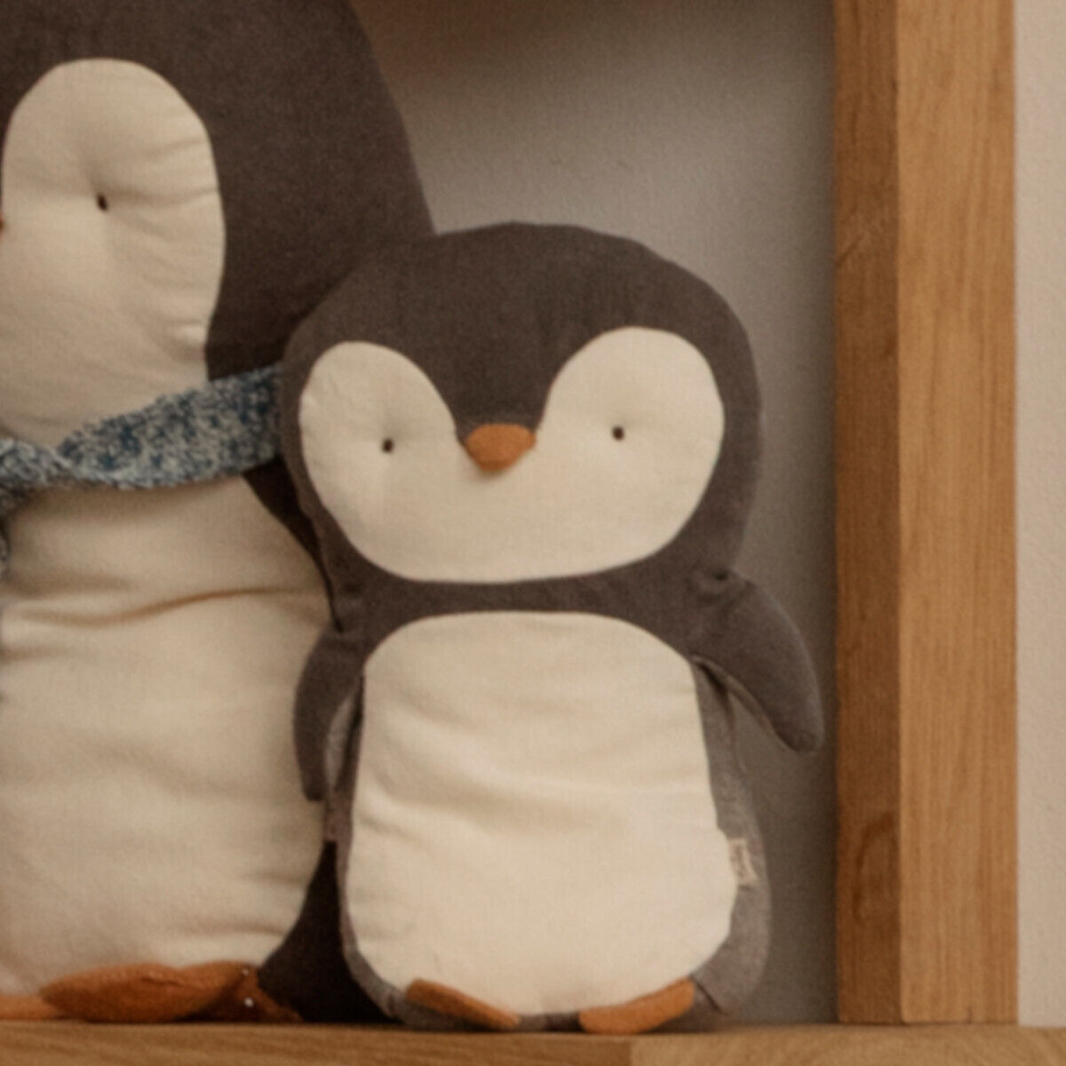 Zwei Kuscheltiere in Form von Pinguinen stehen nebeneinander. Die Pinguine sind grau und weiß, mit orangefarbenen Schnäbeln und Füßen. Der größere Pinguin trägt einen blauen Schal.