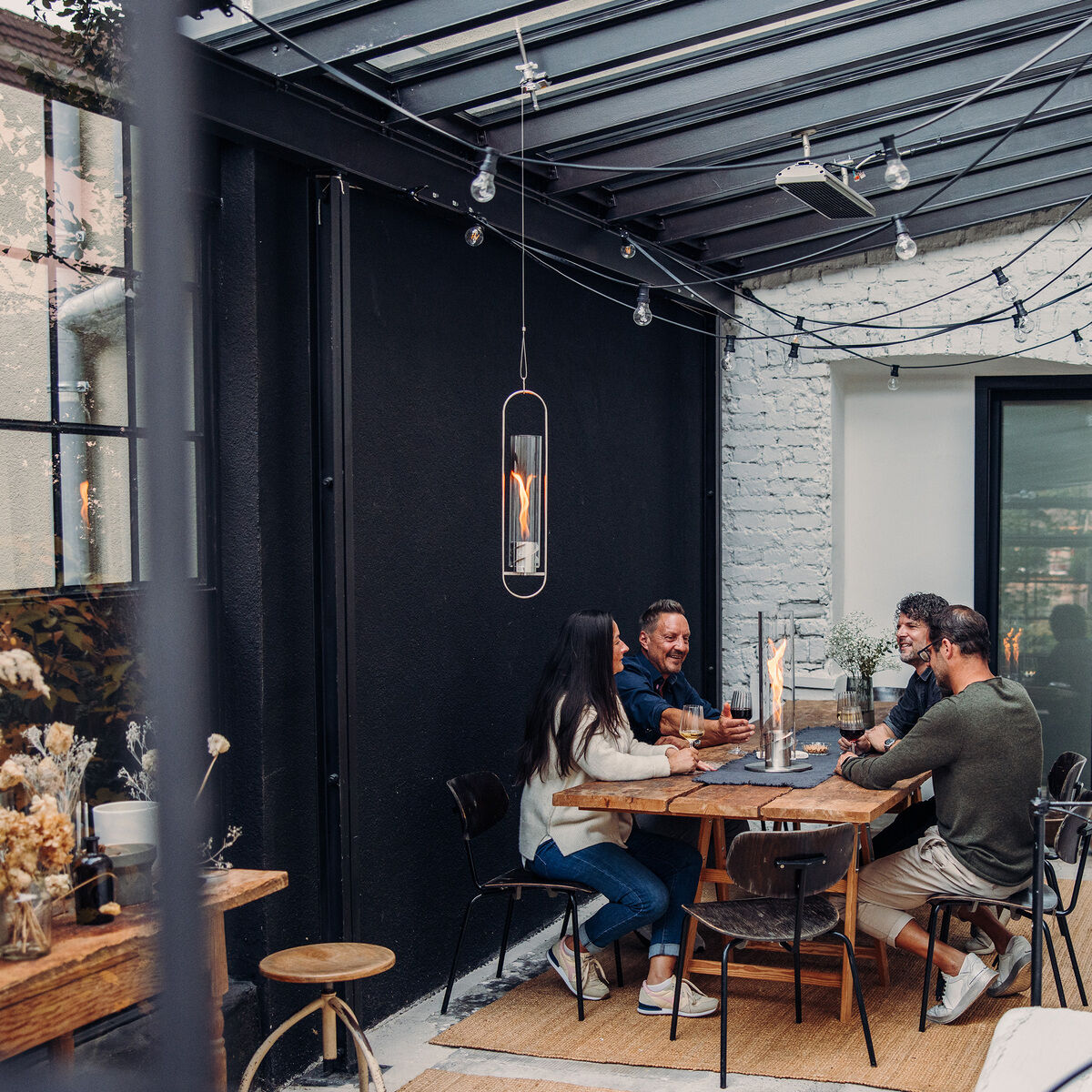 Mehrere Personen sitzen um einen gedeckten Tisch auf einer Terrasse, die mit dem SPIN Hängesystem beleuchtet wird. Die Atmosphäre ist gemütlich, mit Lichterketten und Dekoration überall. Das Design ist modern und stilvoll.