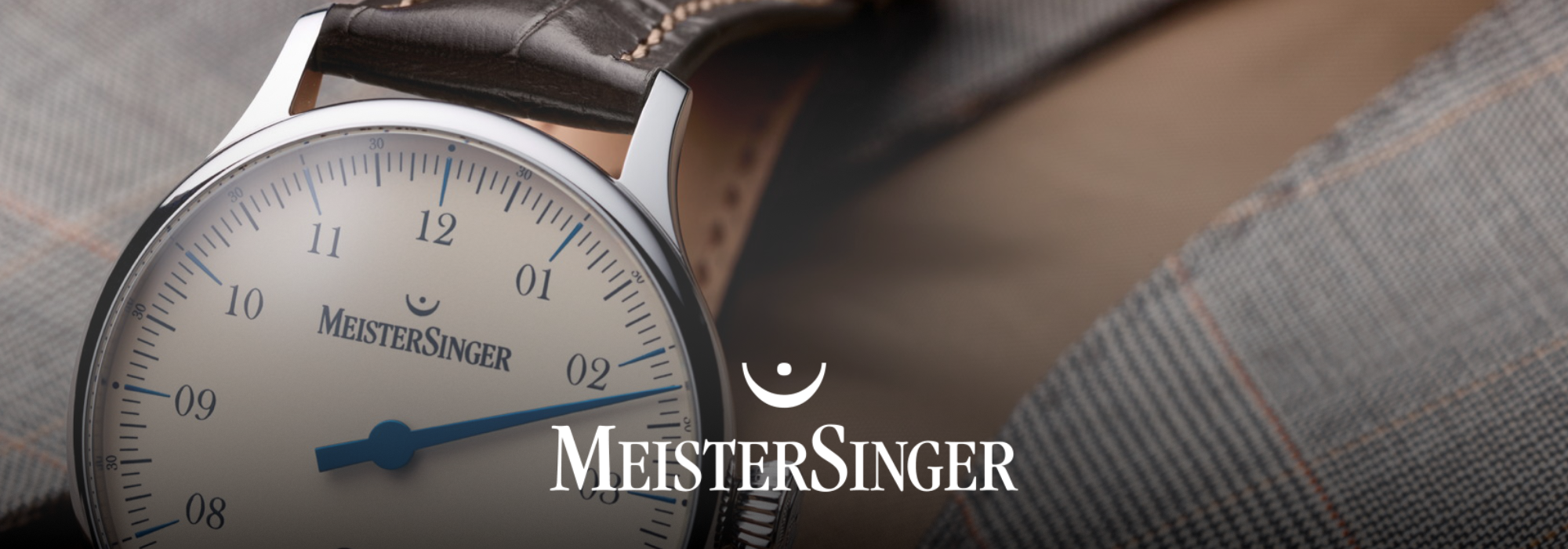 Nahaufnahme einer MeisterSinger-Uhr an einem braunen Lederarmband mit weißem Zifferblatt und Stundenmarkierungen vor einem Hintergrund aus strukturiertem Stoff.