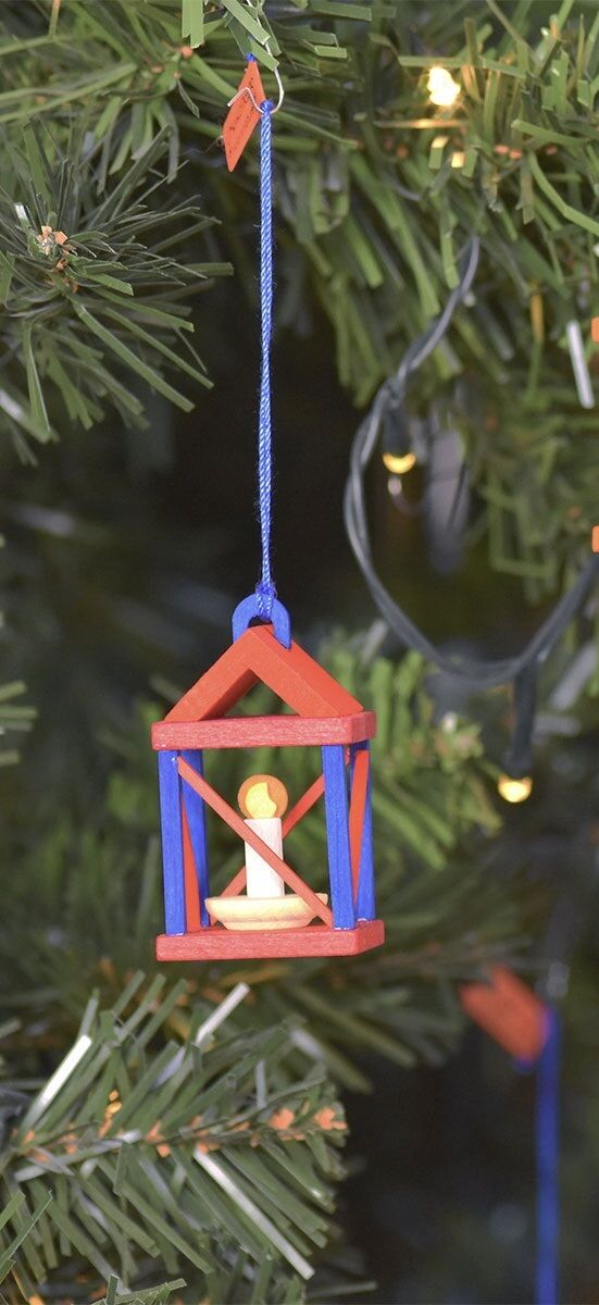 Ein rotes und blaues handgemachtes Holzornament in Form einer Laterne mit einer weißen Kerze in der Mitte, hängt an einem grünen Weihnachtsbaum.
