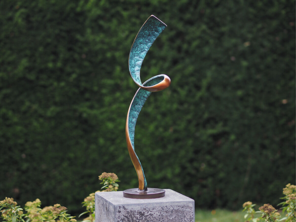 Fotografie einer abstrakten Gartenskulptur aus Metall, die sich auf einer grauen Basis befindet. Die Skulptur hat geschwungene Formen in den Farben Grün und Bronze. Der Hintergrund ist grün, vermutlich Hecken oder Bäume, was auf einen Garten hindeutet.
