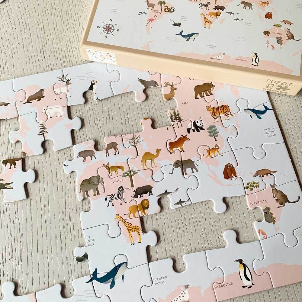 Ein teilweise zusammengesetztes Puzzle mit Tiermotiven auf einer Weltkarte, das auf einem hellen Holztisch liegt. Die Packung des Puzzles steht daneben und zeigt die vollständige Abbildung mit verschiedenen Tieren wie Tiger, Pinguin, Wal, Giraffe, und Panda. Die Farben des Puzzles sind vorwiegend beige und pastellfarben.