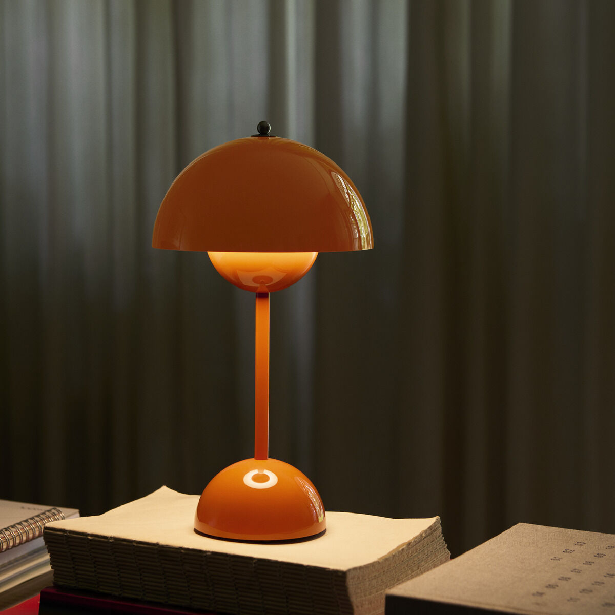 Im Mittelpunkt steht die orangefarbene Flowerpot VP9 Tischleuchte von &Tradition auf einem Stapel Bücher mit beiger Oberfläche. Die Retro-Design-Lampe leuchtet angenehm warm und diffuses Licht fällt auf die Bücher. Im Hintergrund verdecken lange, graugrüne Vorhänge das Fenster.