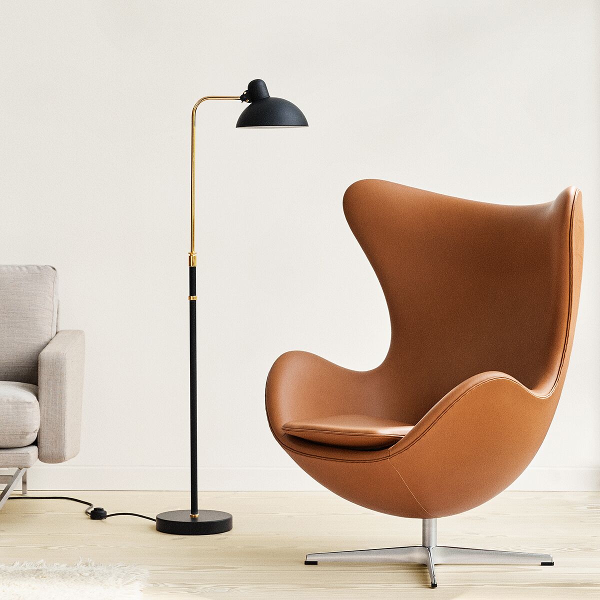 Die Stehlampe »KAISER IDELL« matt von Fritz Hansen steht neben einem hellbraunen Design-Ledersessel im bekannten Egg Chair-Stil und einem grauen Sofa. Die Lampe ist aus Stahlblech und Chrom, schwarz-matt mit messingfarbenem Standrohr und rundem Fuß. Im Hintergrund ist eine schlichte, weiße Wand.