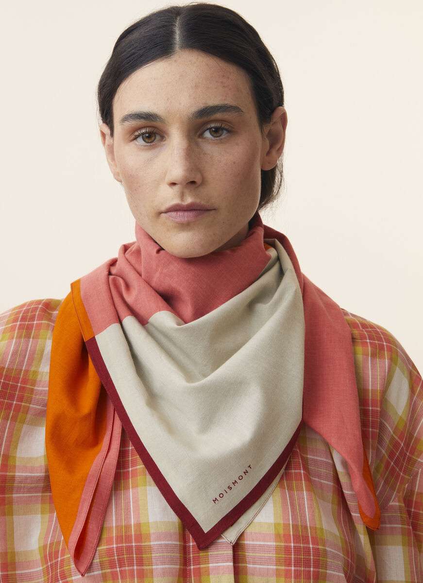 Frau trägt ein Baumwolltuch mit rosa, cremefarbenen, orange und roten Tönen. Das Tuch ist modisch drapiert und ergänzt ein kariertes Oberteil, was einen stilvollen und trendbewussten Look ergibt.
