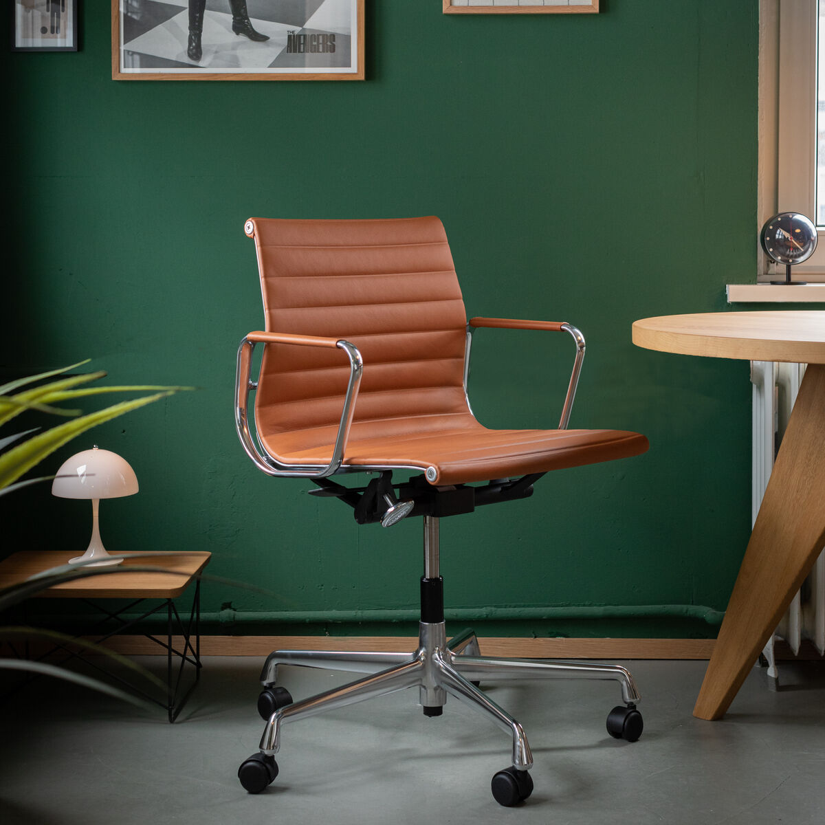 Vitra Alu Chair EA 118, ein polierter, ergonomischer Bürostuhl aus braunem Leder und Aluminiumrahmen, steht vor einer grünen Wand mit gerahmten Bildern. Ein modernes Büro-Setup mit einem Schreibtisch, einer Lampe und Pflanzen ist sichtbar.