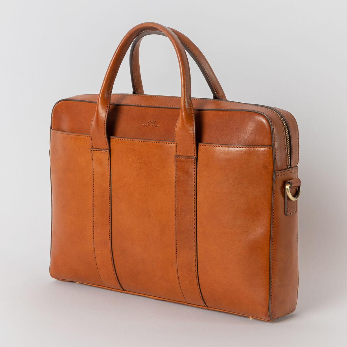 Braune Ledertasche des Modells 'Harvey' vor grauem Hintergrund. Die Tasche ist eckig mit zwei Griffen und einem goldfarbenen Reißverschluss.