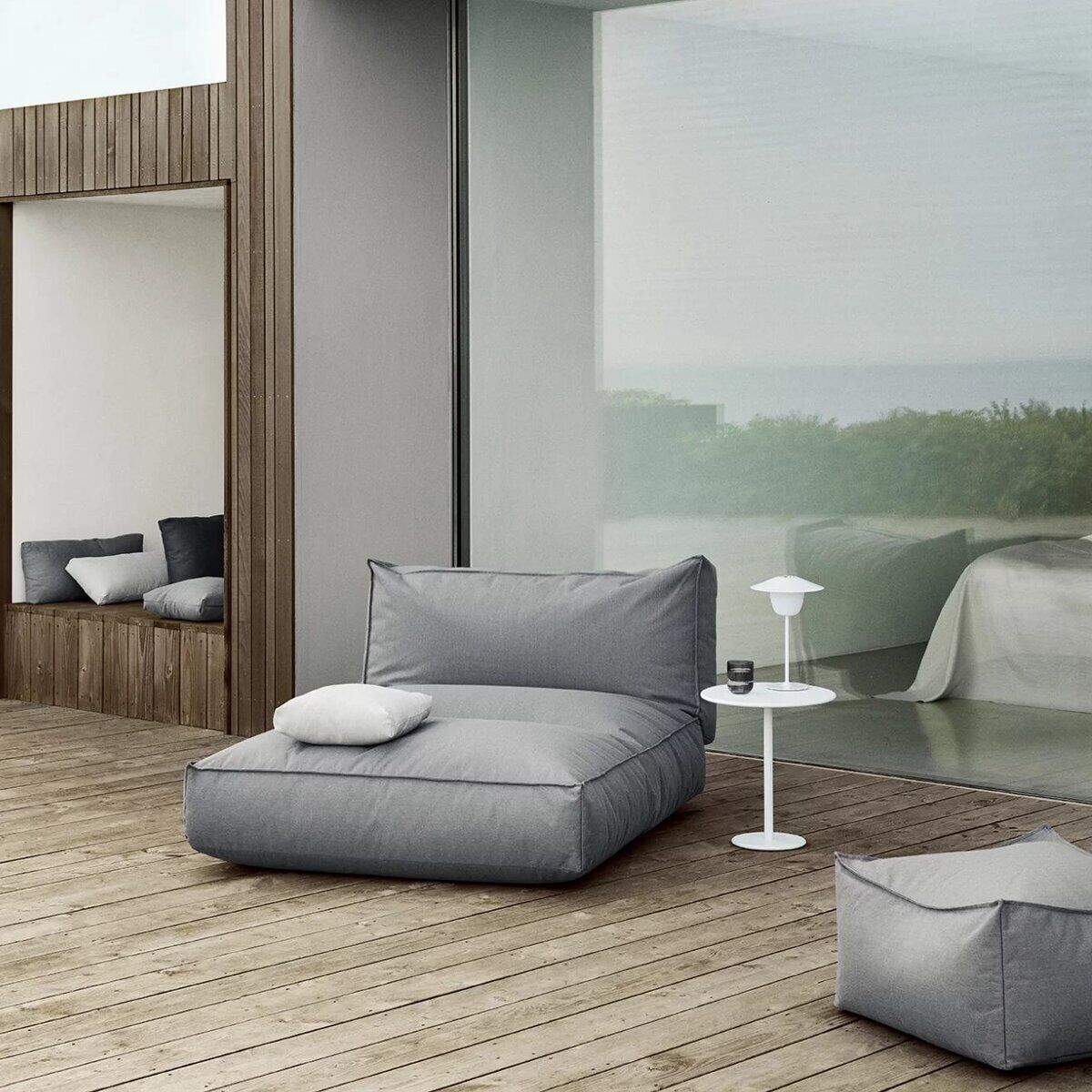 Ein modernes Outdoor-Bett »STAY« mit einer Größe von 80 x 190 cm ist auf einer stilvollen Terrasse mit Holzboden abgebildet. Das Bett hat eine graue Polsterung und ist von einem kleinen weißen Beistelltisch und einem kleinen grauen Pouf umgeben. Im Hintergrund sind moderne architektonische Elemente und eine Glaswand sichtbar.