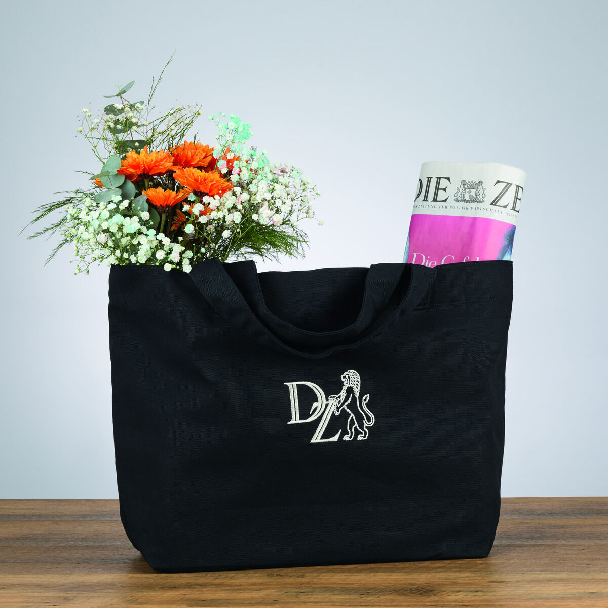 Eine schwarze Canvas-Tasche mit dem ZEIT-Logo, gefüllt mit einem Blumenstrauß und einer Zeitung, auf einem Holztisch vor einem hellen Hintergrund.