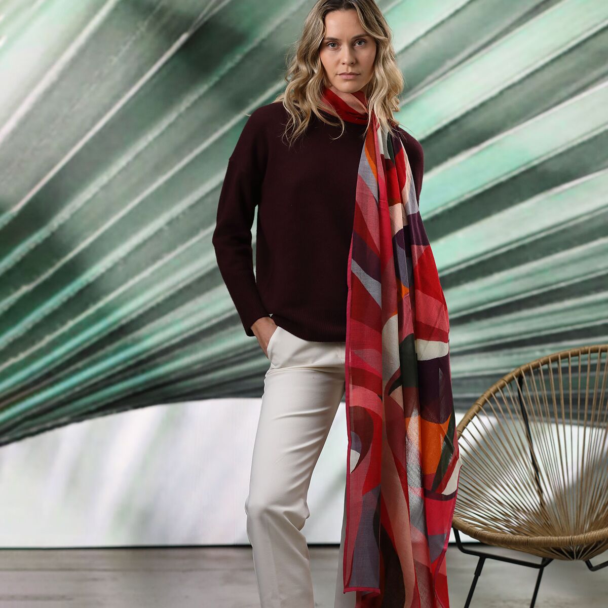 Frau mit langem, gemustertem Wollschal in Rottönen trägt einen dunkelroten Pullover und weiße Hose vor modernem Hintergrund. Der Schal zeigt ein farbenfrohes, geometrisches Design.