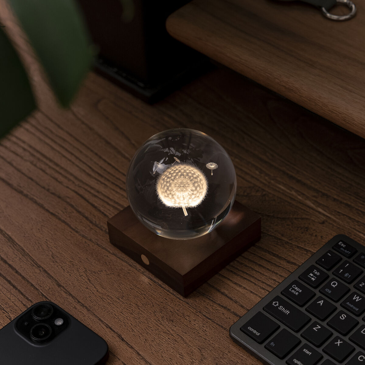 Auf einem Holzschreibtisch steht die elegante Lampe »Amber Light« von Gingko Design Ltd mit einer klaren, kristallenen Glaskugel und einem Sockel aus dunklem Walnussholz. Die Leuchte strahlt warmes Licht und schafft ein stilvolles, gemütliches Ambiente zwischen Tastatur und Smartphone.