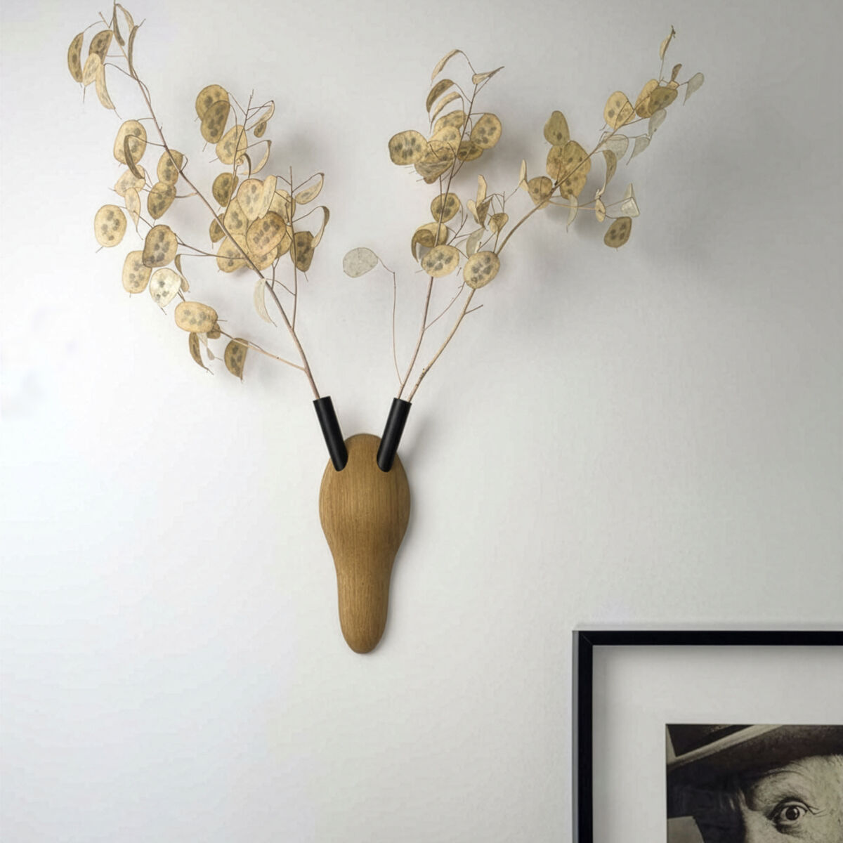 Detailreiche Wanddeko »RESPECT« von finbeaux aus Esche massiv: Hirschgeweih-Optik mit filigranen Zweigen, mittig Holzteil, zwei schwarze Stahlelemente, darunter ein schwarz gerahmtes Bild mit Porträt, alles auf hellem Hintergrund, modernes Ambiente.