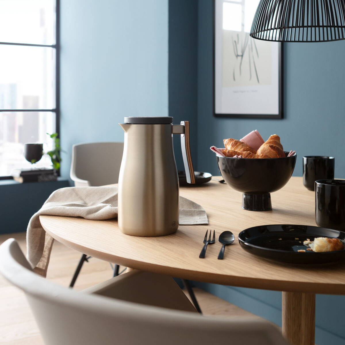Eine moderne Edelstahl-Isolierkanne steht auf einem Holzfrühstückstisch, umgeben von schwarzen Kaffeetassen, Besteck und einem Korb mit Croissants. Der Raum hat blaue Wände und ein Fenster mit Blick auf die Stadt.
