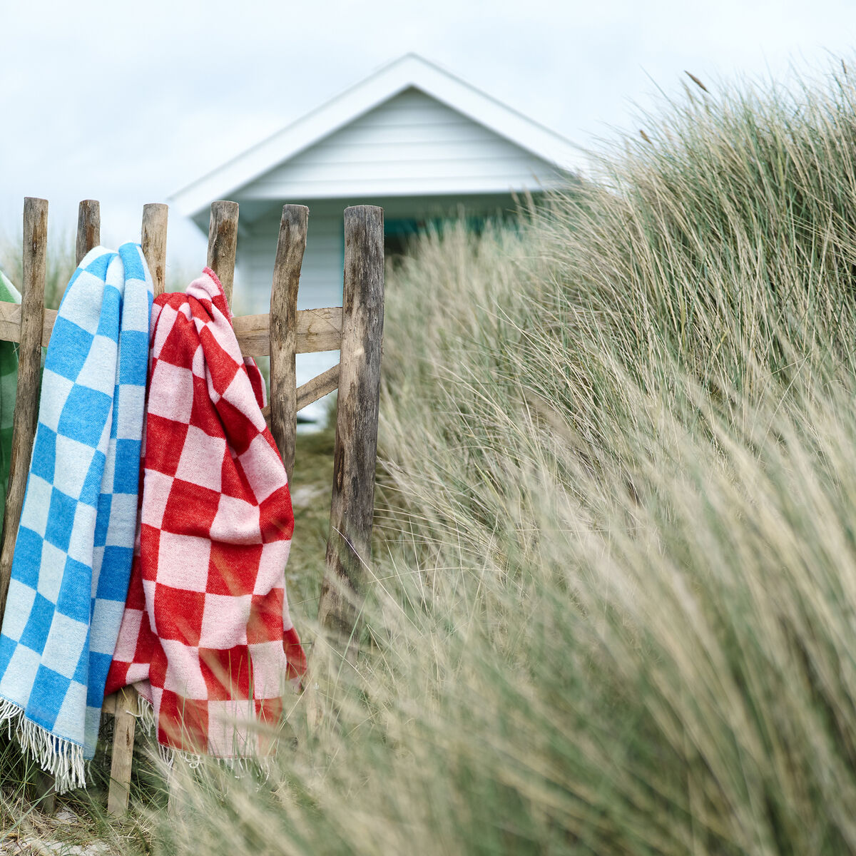 Drei Decken in Karomuster – grün-weiß, blau-weiß und rot-weiß – hängen an einem Holzzaun. Im Hintergrund sind grasbewachsene Dünen und eine Strandhütte sichtbar, unter einem bewölkten Himmel.