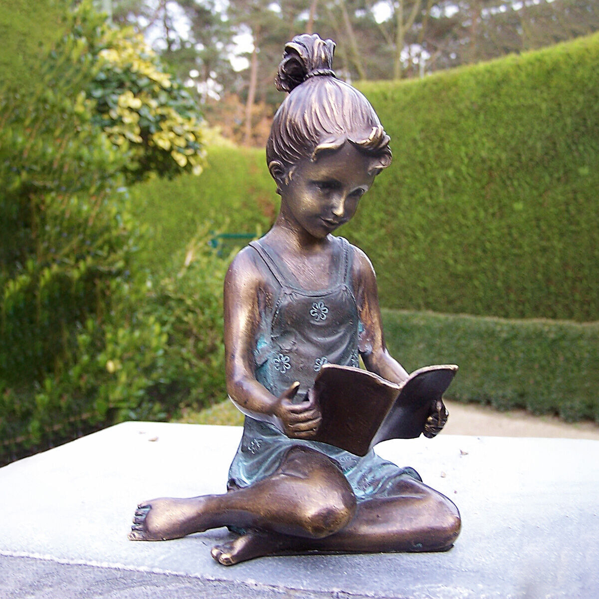 Eine Bronzeskulptur eines Mädchens, das in einem Garten sitzt und ein Buch liest. Die Skulptur zeigt detaillierte Merkmale und steht vor einem Hintergrund mit grüner Hecke und Pflanzen.