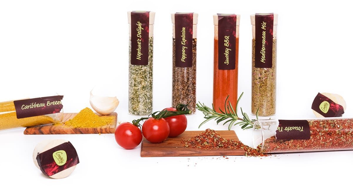 Ein Set von fünf verschiedenfarbigen, länglichen Gewürzdosen mit Etiketten, umgeben von Tomaten, Kräutern, Knoblauch und Gewürzen auf einem weißen Hintergrund.