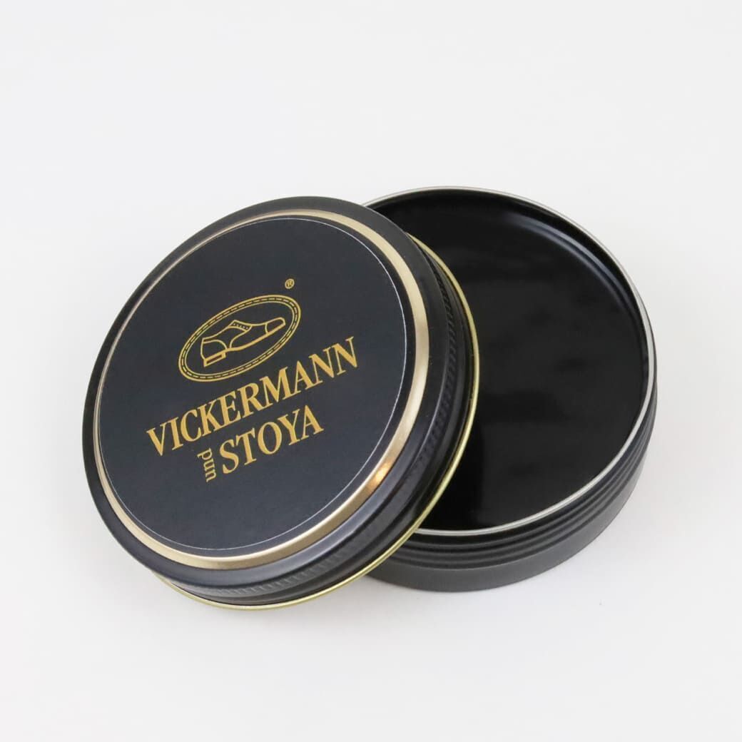 Eine runde Dose mit schwarzer Lederpflegecreme von Vickermann und Stoya. Der Deckel ist schwarz mit goldener Beschriftung und Rand. Die Dose ist offen, auf einer weißen Oberfläche fotografiert.