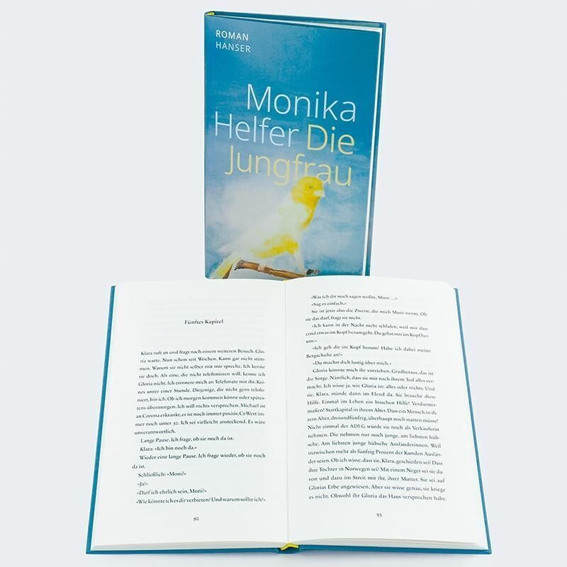 Das Bild zeigt den Roman 'Die Jungfrau' von Monika Helfer. Das Buch steht aufrecht im Hintergrund, während ein geöffnetes Exemplar im Vordergrund liegt, das Innere zeigt einige Textseiten. Das Cover ist hauptsächlich blau mit gelbem Text.