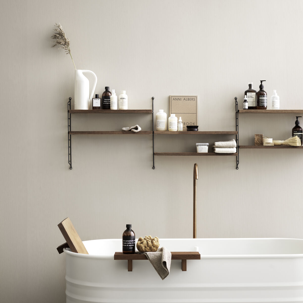 Ein minimalistisches Wandregal namens »String Pocket Regal« mit Holzlatten und schwarzen Metallstützen, darauf befinden sich verschiedene Pflegeprodukte. Im Vordergrund steht eine freistehende weiße Badewanne, auf deren Rand mehrere Badezimmerutensilien platziert sind.