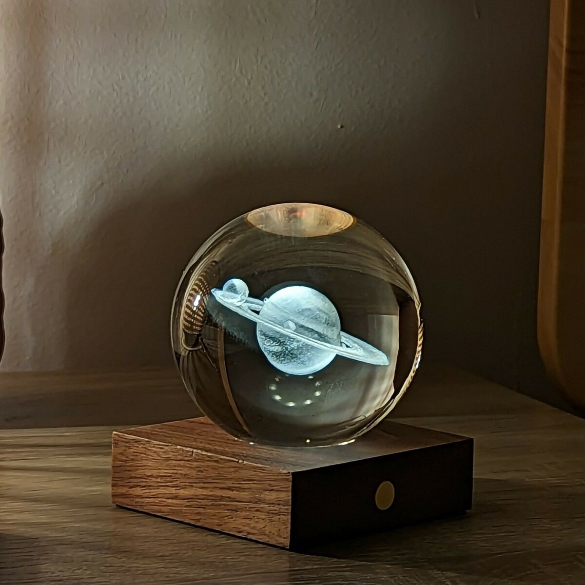 Eine moderne Lampe mit einer kugelförmigen Kristallkugel, die ein beleuchtetes Saturnmodell zeigt, steht auf einer eleganten Holzplatte auf einem Tisch in einem stilvoll eingerichteten Raum.