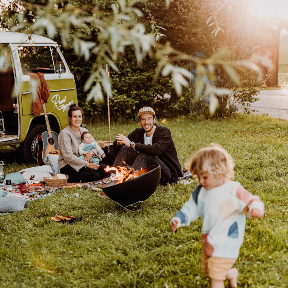 Ein Picknick im Freien mit einer Familie, einem grünen Camper, einer Gitarre und einem Feuerkorb. Ein kleines Kind nähert sich dem Feuer, während die Eltern auf einer Decke mit ihrem Baby sitzen.