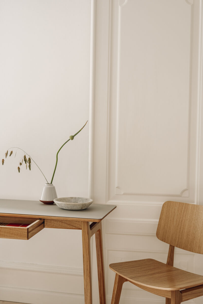 Ein minimalistischer Schreibtisch aus Eiche namens »Freya« steht vor einer cremeweißen, verzierten Wand. Auf dem Schreibtisch befinden sich eine schlichte Vase mit Grasdecor und eine Keramikschale. Ein passender Holzstuhl steht daneben.