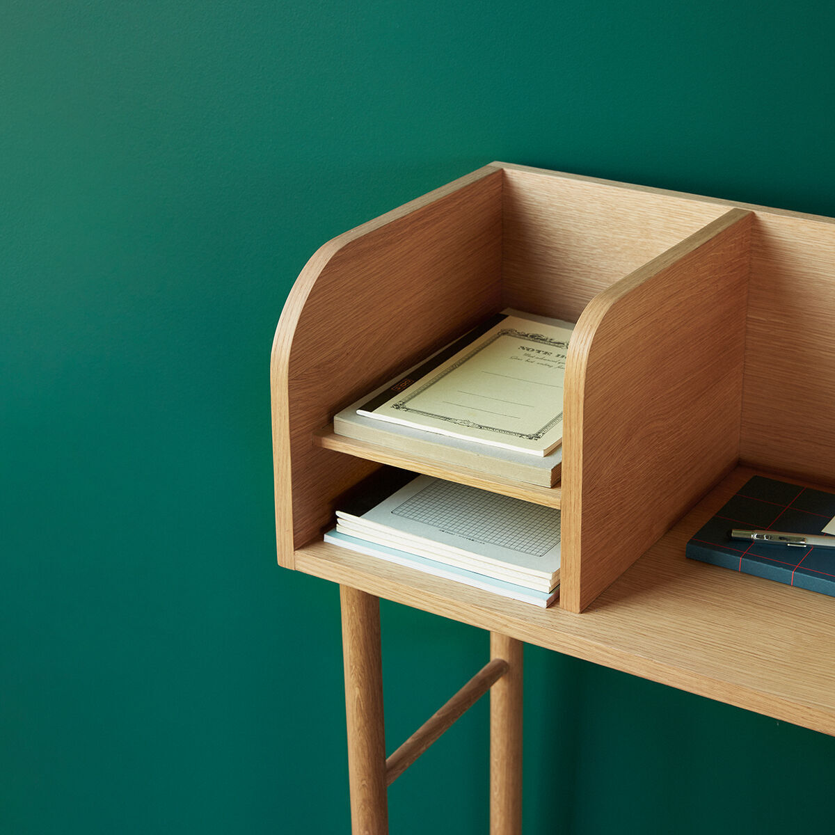 Schreibtisch aus Eichenholz mit zwei getrennten Ablagebereichen; grüne Wand im Hintergrund und Bücher bzw. Notizbücher auf der Tischplatte.