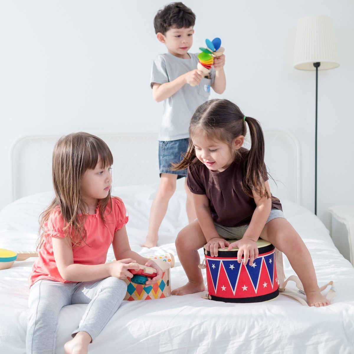 Drei Kinder spielen zusammen auf einem Bett mit verschiedenen Musikinstrumenten, darunter eine große Trommel mit einem roten Korpus und blauen sowie weißen Details.
