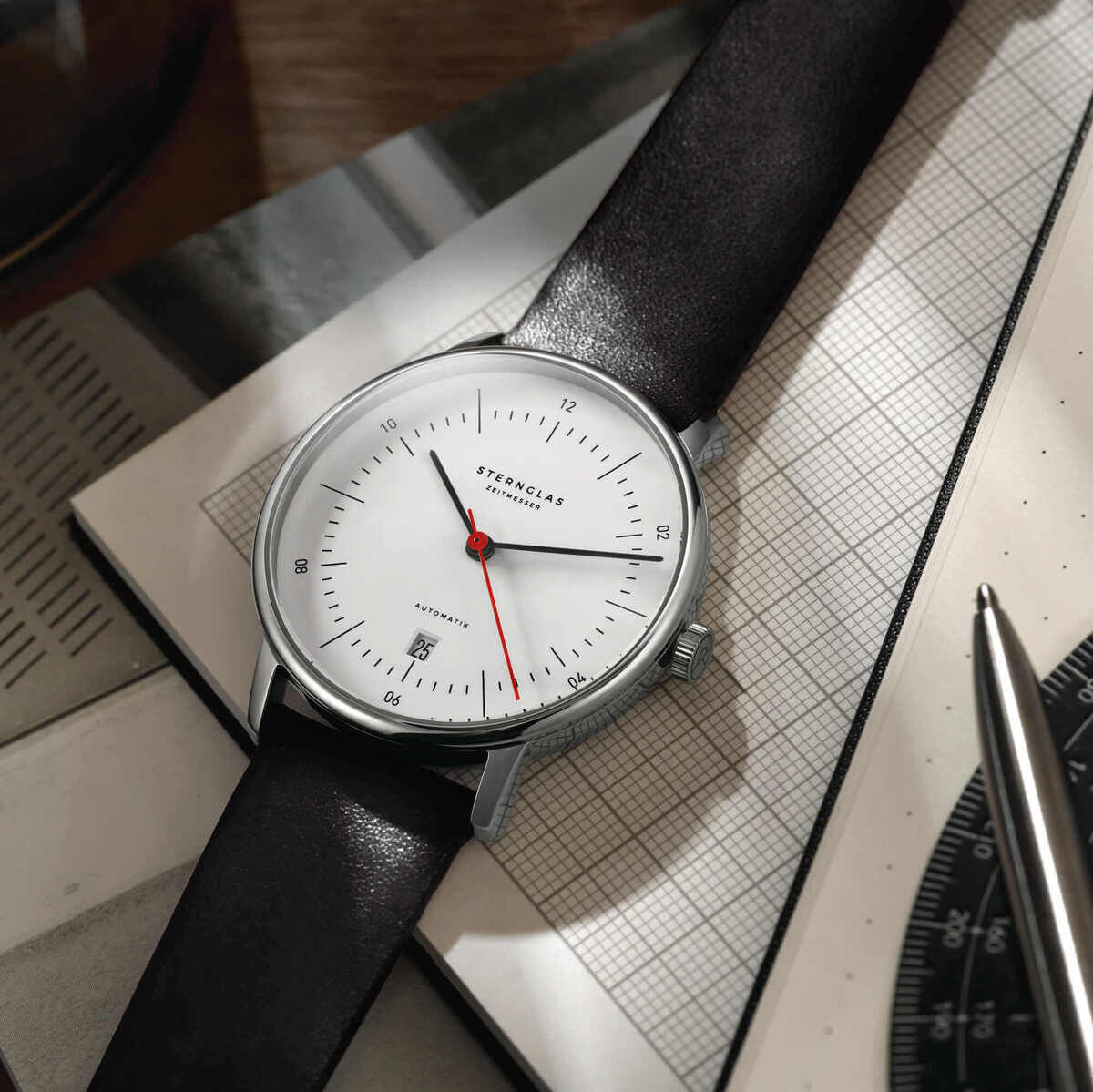 Eine elegante Herrenuhr der ZEIT-Sonderedition »Naos Automatik« mit einem schwarzen Lederarmband und einem minimalistischen Zifferblatt. Die Uhr liegt auf einem Schreibtisch mit einer Schreibunterlage und einem Stift.