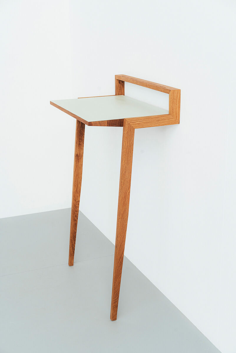 Ein minimalistisches Stehpult aus Holz mit einer weißen Arbeitsfläche. Es hat zwei schmale Beine und ist an eine Wand gelehnt. Der Stil ist modern und platzsparend.