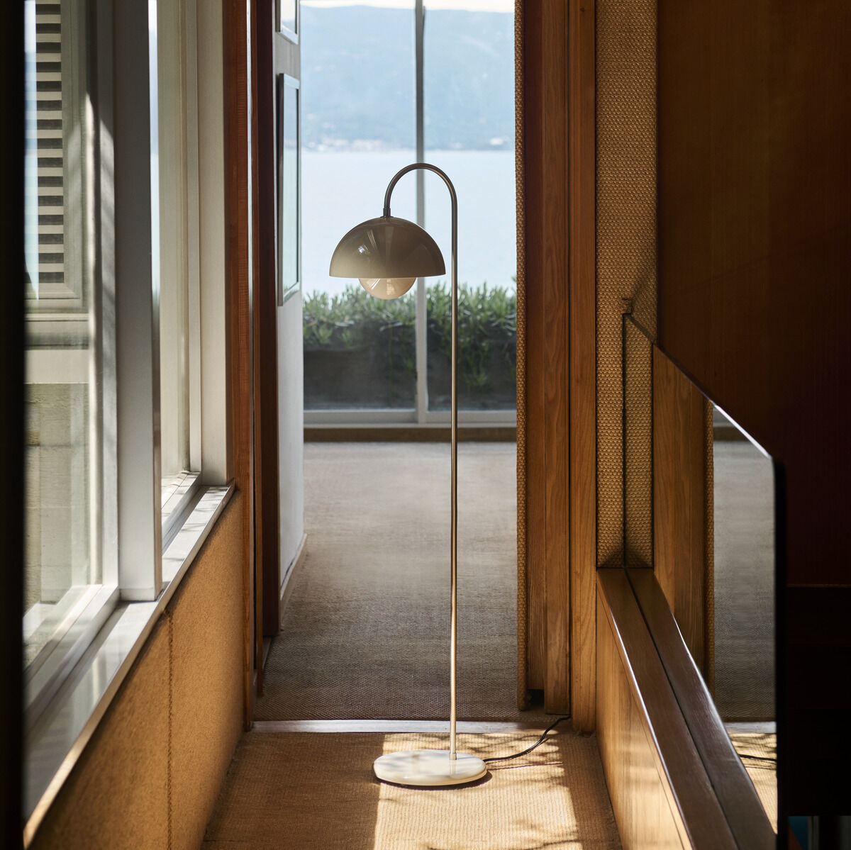 Eine stilvolle Standleuchte im minimalistischen Design, die in einem modern eingerichteten Flur mit Holzelementen steht. Durch ein großes Fenster im Hintergrund fällt Tageslicht ein, das den Flur beleuchtet. Draußen ist die Aussicht auf einen ruhigen See zu sehen.