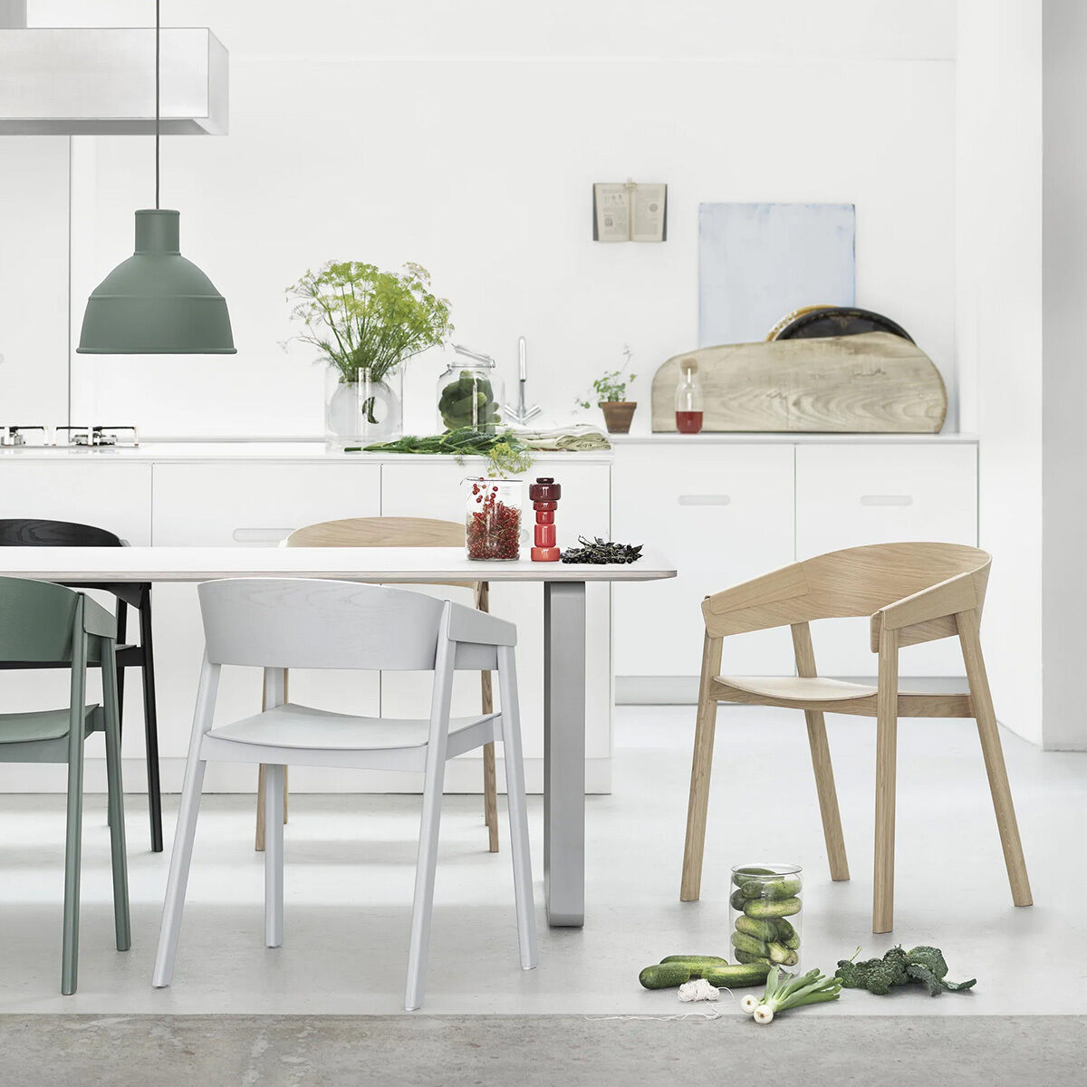 Helles, modernes Esszimmer mit weißen Wänden, minimalistischer Küche und drei Cover Stühlen »Eiche« von Muuto in verschiedenen Farben. Auf dem Tisch und Boden liegen Gemüse und Kräuter. Schlichte skandinavische Ästhetik mit natürlichem Lichteinfall und klaren Linien.