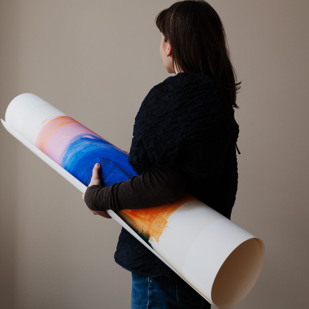 Eine Person hält ein gerahmtes, abstraktes Kunstwerk von Berit Mogensen Lopez mit dem Titel 'Improvise'. Das Bild zeigt einen Farbverlauf in Blau, Rosa und Orange auf weißem Hintergrund. Die Szene wirkt modern und minimalistisch.