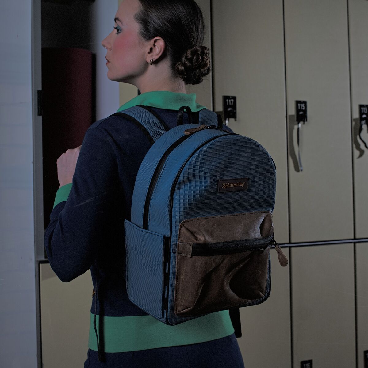 Ein Rucksack »ZEIT Gymnast« aus Stoff und Leder, getragen von einer Person vor einer Schrankreihe. Der Rucksack hat mehrere Fächer und einen Reißverschluss; die Farben sind hauptsächlich Blau und Braun.