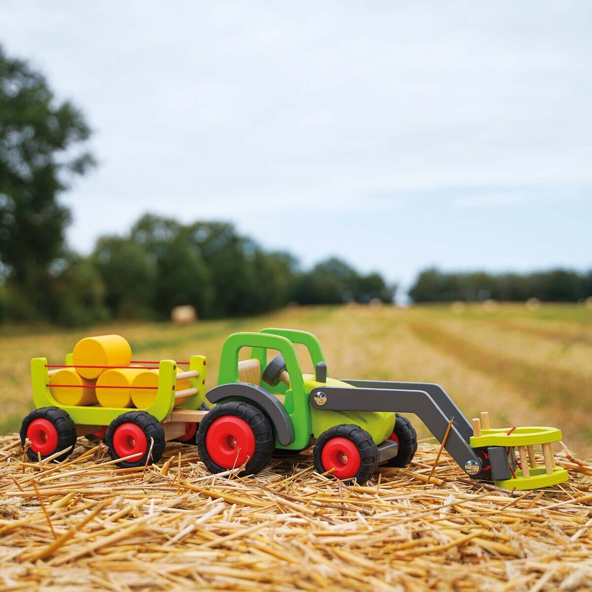 Ein Holz-Frontlader-Traktor mit Heuwagen steht auf einem Strohfeld. Der grüne Traktor mit roten Rädern zieht einen Anhänger, auf dem gelbe Holzballen liegen. Im Hintergrund sind Bäume und ein weites Erntefeld zu sehen, unter einem bewölkten Himmel.