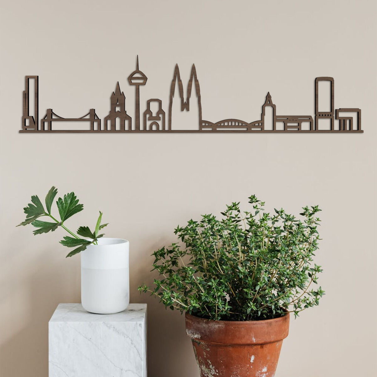 Dekoration aus Holz an der Wand dargestellt, die die Skyline von Köln darstellt. Die Silhouette umfasst bekannte Gebäude der Stadt. Im Vordergrund sind eine grüne Pflanze und ein weißes Gefäß auf einem Sockel sichtbar.