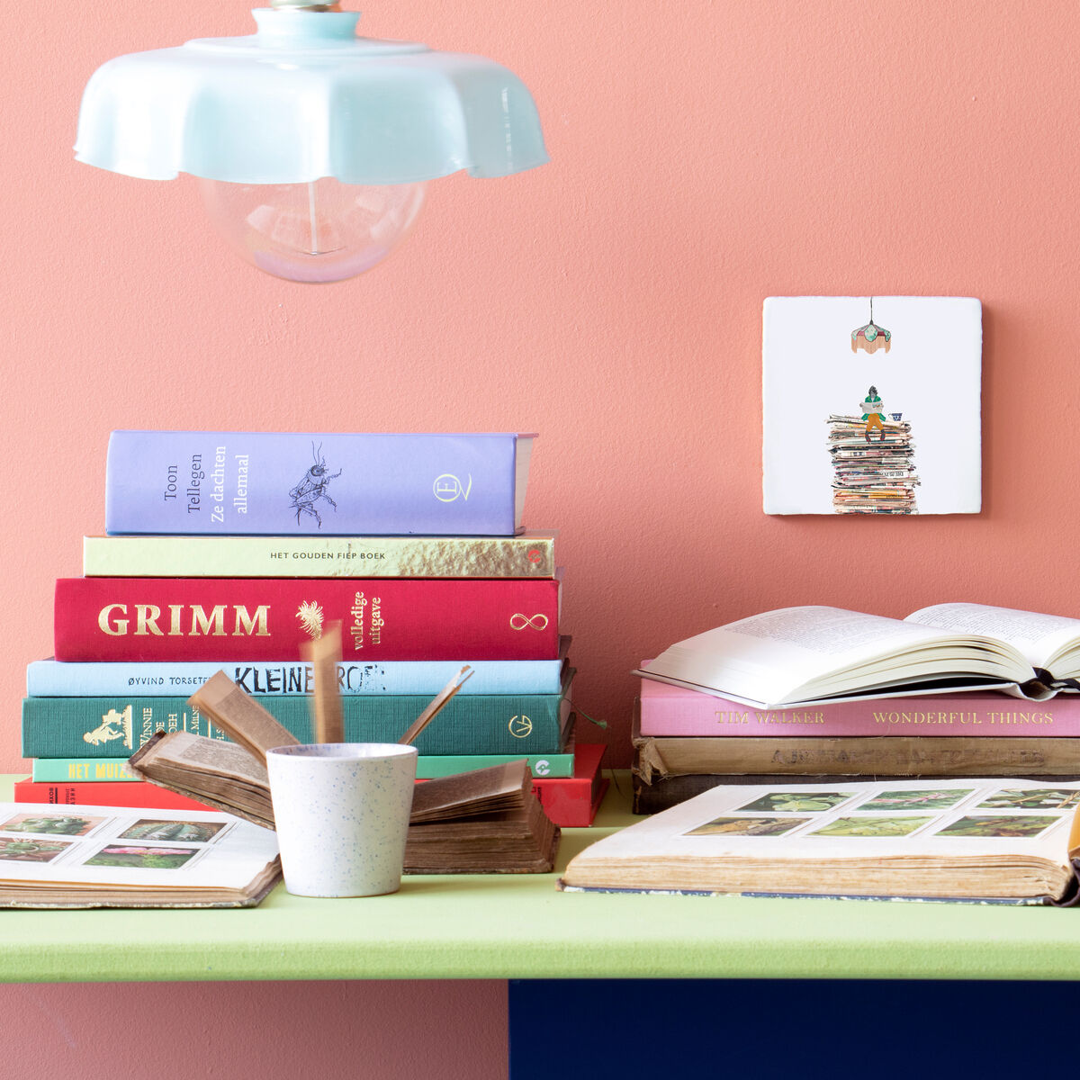 Die Keramikfliese »DIE ZEIT« ist an einer rosa Wand über einem Schreibtisch mit Büchern, Stiften und einer Lampe befestigt. Die Fliese zeigt eine Illustration eines Bücherstapels.