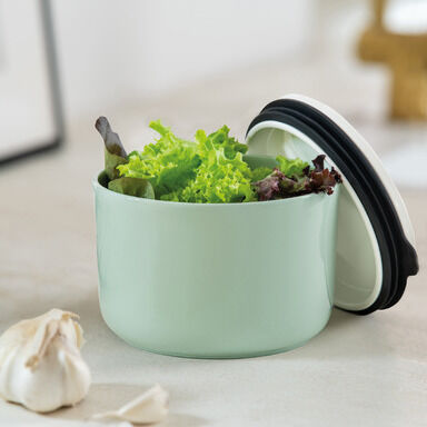 Grüne runde Lunchbox mit Deckel in weiß und schwarz, gefüllt mit frischem grünem Salat, auf einer weißen Oberfläche. Keine anderen Gegenstände bis auf Knoblauchzehen im Bildhintergrund erkennbar.