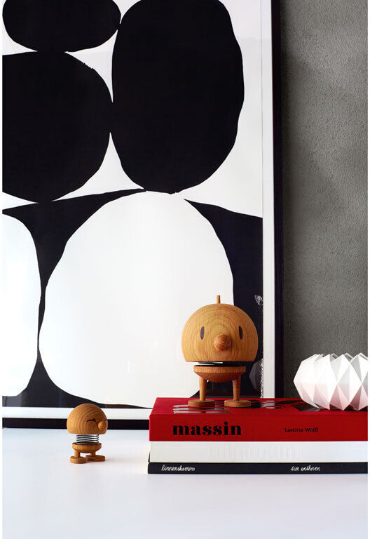 Das Bild zeigt zwei Hoptimist Bumble Figuren aus Eiche auf einem weißen Hintergrund. Eine Figur steht auf einem Stapel von Büchern, und im Hintergrund ist ein minimalistisches Kunstwerk mit schwarzen und weißen Formen zu sehen.