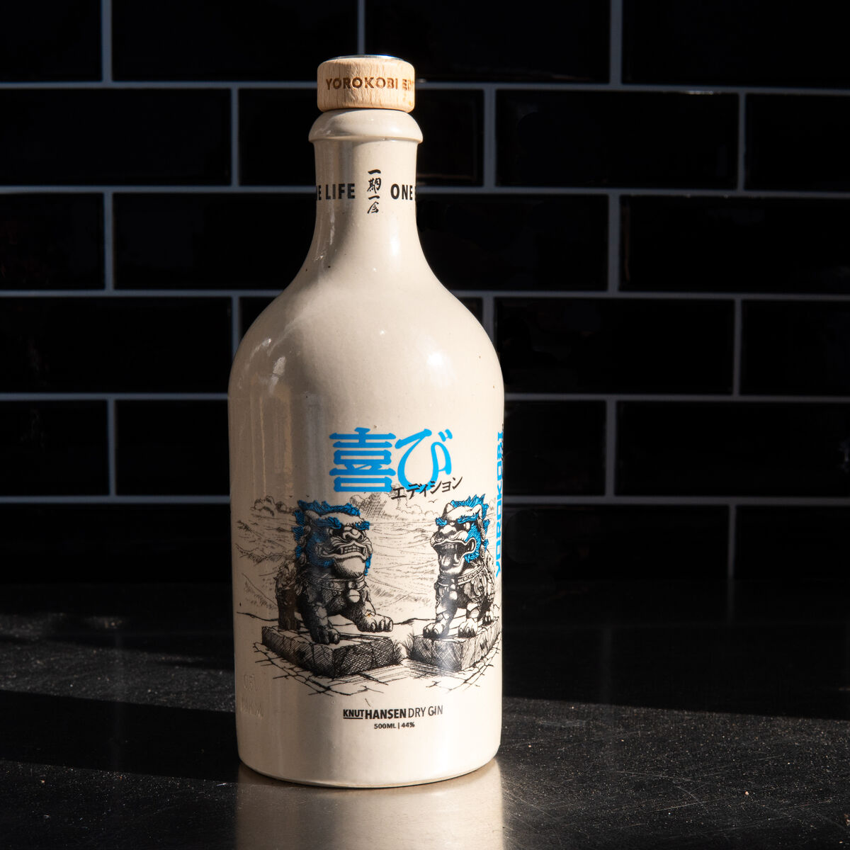 Die Knut Hansen Sonderedition 2024 YOROKOBI ist eine weiß-beige Gin-Flasche mit einem japanisch inspirierten Design. Das Bild zeigt zwei detaillierte Zeichnungen von Steinfiguren sowie blauen Text auf der Vorderseite der Flasche. Sie steht auf einer reflektierenden Oberfläche vor einem dunklen, gekachelten Hintergrund.