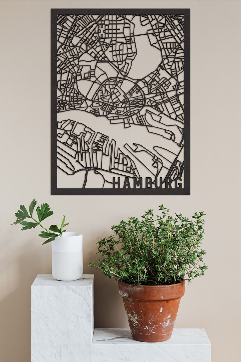 Eine minimalistische Holzkunstkarte von Hamburg an einer beigefarbenen Wand. Die Karte zeigt detailliert die Stadtstruktur. Darunter steht eine grüne Pflanze in einem Terrakottatopf und eine weiße Vase auf einem weißen Podest.