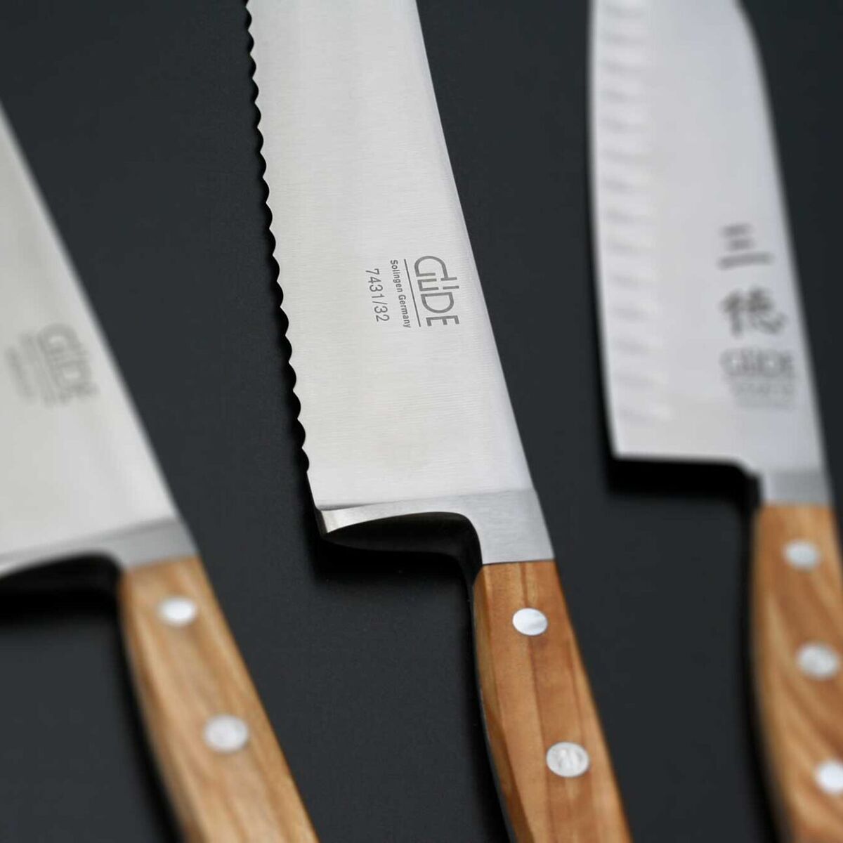 Drei Küchenmesser mit scharfen Edelstahlklingen und ergonomischen Holzgriffen liegen nebeneinander auf einer dunklen Oberfläche. Das Brot- und Santokumesser sind gut erkennbar. Die Messer sind für den Küchengebrauch und als Geschenk geeignet.